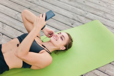Spor giyim bölümündeki neşeli genç bayan müzik dinliyor ve ahşap terasta minderde yatarken akıllı telefonu elinde tutuyor.