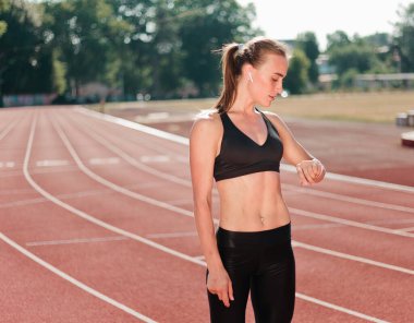 Spor giyimli neşeli bayan koşucu, açık havada kırmızı kaplamalı stadyumdaki akıllı saate bakıyor.