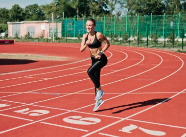 Spor kıyafetleriyle koşan neşeli kadın koşucu stadyum pistinde kırmızı kaplamayla koşuyor.
