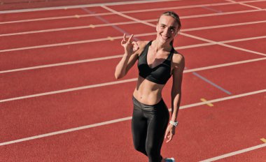 Neşeli kadın koşucu iyi bir jest yapıyor ve kırmızı kaplı stadyumda gülümsüyor.