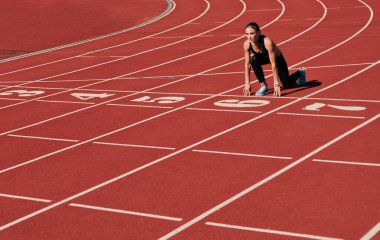 Spor giyimli genç bir kadın, parlak güneşli bir günde, stadyum pistinde koşuya çıkmaya hazırlanıyor.