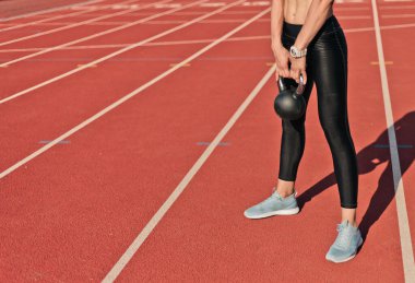 Güçlü zayıf kadın, güneşli bir günde stadyumda kırmızı kaplamalı kettlebell ile güç egzersizi yapıyor.