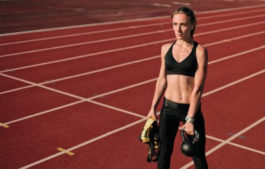 Spor kıyafetleri içinde genç çekici bir bayan sporcu elinde spor ekipmanı (kettlebell ve fit strap) parlak güneşli bir günde elinde kırmızı kaplama ile stadyum pistinde.