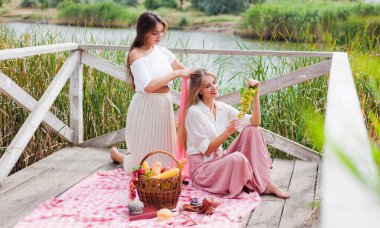 İki neşeli genç kadın yaz günü dışarıda piknik yapıyorlar. Eski moda elbiseli iki kız arkadaş göl kıyısında birlikte vakit geçiriyorlar..