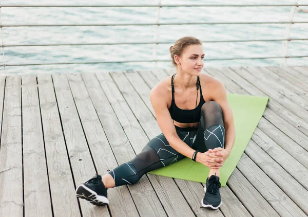 身穿运动服的年轻肌肉发达的女人坐在沙滩平台的垫子上休息 健康生活方式概念 — 图库照片