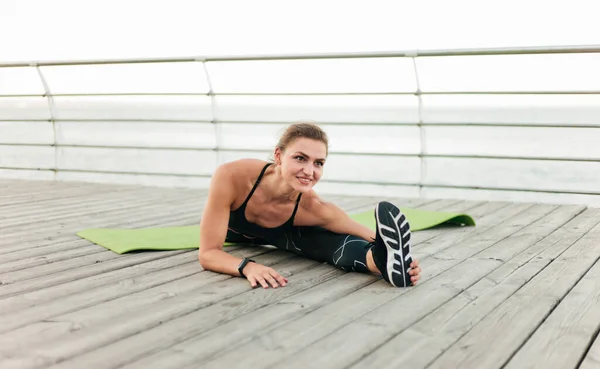身穿运动服的肌肉发达的女运动员坐在海滨露台上 双腿伸展 户外运动 — 图库照片
