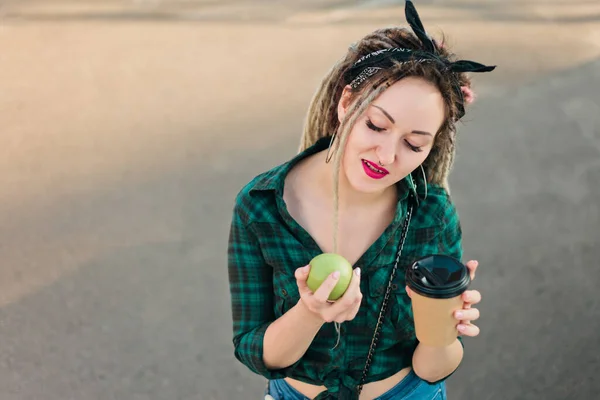 Attractive woman with an informal haircut dreadlocks hesitates before choosing an apple or coffee outdoors