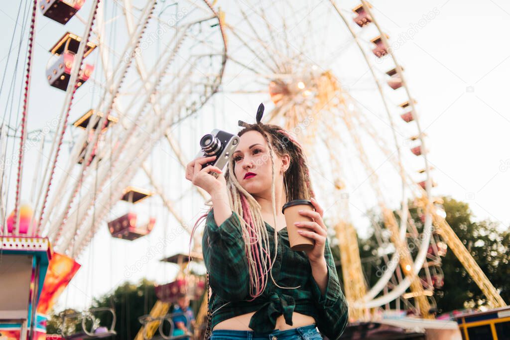 An informal young woman with dreadlocks takes pictures with retro camera in amusement park.