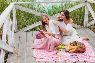 İki neşeli genç kadın yaz günü dışarıda piknik yapıyorlar. Eski moda elbiseli iki kız arkadaş göl kıyısında birlikte vakit geçiriyorlar..