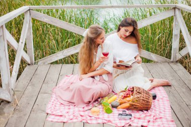 İki neşeli genç kadın yaz günü dışarıda piknik yapıyorlar. Eski moda elbiseli iki kız arkadaş göl kıyısında birlikte vakit geçiriyorlar..