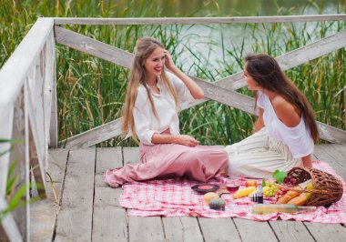 İki neşeli genç kadın yaz günü dışarıda piknik yapıyorlar. Eski moda elbiseli iki kız arkadaş göl kıyısında birlikte vakit geçiriyorlar..