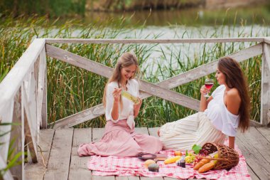 İki neşeli genç kadın yaz günü dışarıda piknik yapıyorlar. Eski moda elbiseli iki kız arkadaş göl kıyısında birlikte vakit geçiriyorlar..