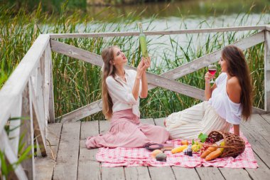 İki neşeli genç kadın yaz günü dışarıda piknik yapıyorlar. Eski moda elbiseli iki kız arkadaş göl kıyısında birlikte vakit geçiriyorlar..