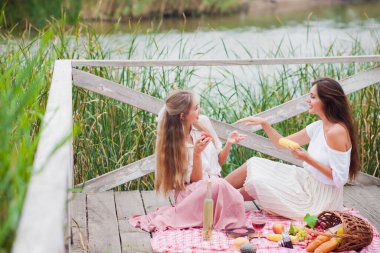 İki neşeli genç kadın yaz günü dışarıda piknik yapıyorlar. Eski moda elbiseli iki kız arkadaş göl kıyısında birlikte vakit geçiriyorlar..