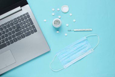 Tıbbi geçmişi var. Modern bir doktorun çalışma alanı. Laptop, haplar, gazlı bez maskesi, mavi arka planda termometre. Üst Manzara. Düz yatıyordu
