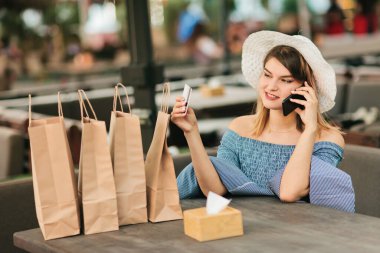İnternetten alışveriş. Neşeli, alışveriş düşkünü bir kadın açık kafede oturmuş telefonla konuşuyor ve elinde banka kartı tutuyor.