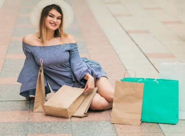Alışveriş konsepti. Şapkalı ve elbiseli neşeli alışverişçi kadın elinde poşetlerle yerde oturuyor.