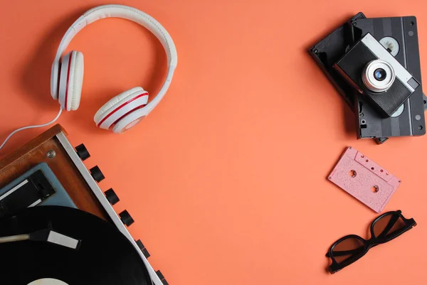 Flat lay retro 80s pop culture objects. Vinyl player, headphones, audio cassette, video tapes, film camera on coral color background. Copy space. Top view