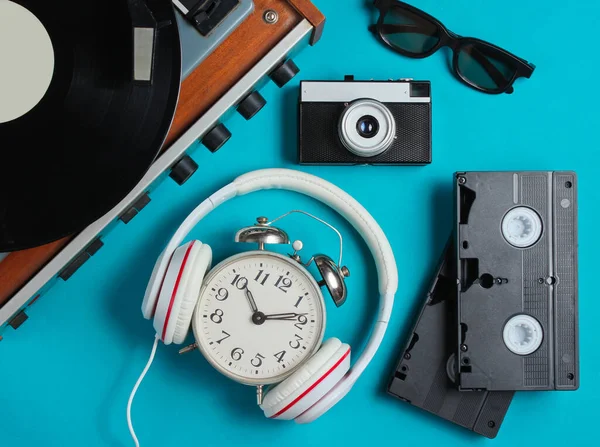 Flat lay retro 80s pop culture objects. Vinyl player, headphones, video tapes, alarm clock, film camera on blue background. Top view