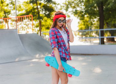 Parlak güneşli bir günde kaykay parkında elinde kaykay tutarken şık giyinmiş neşeli, duygusal genç bir kadın.