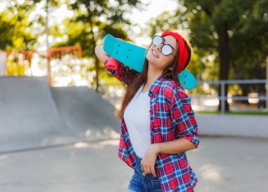 Parlak güneşli bir günde kaykay parkında elinde kaykay tutarken şık giyinmiş neşeli, duygusal genç bir kadın.
