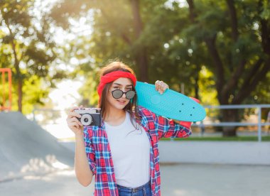 Güneşli bir günde elinde kaykay ve retro kamera tutan şık hippi kız. Gençlik konsepti. Moda düşkünü genç bir kızın yaz hayatı. 