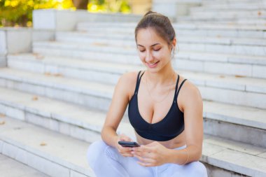 Eğitimden sonra dinlenme kavramı. Spor giyim sektöründe genç bir bayan güneşli bir günde merdivenlerde otururken akıllı telefon kullanır ve kulaklıkla müzik dinler.