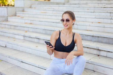 Eğitimden sonra dinlenme kavramı. Spor giyim sektöründe genç bir bayan güneşli bir günde merdivenlerde otururken akıllı telefon kullanır ve kulaklıkla müzik dinler.