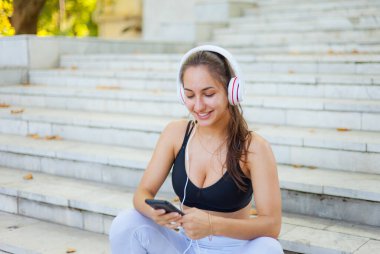 Eğitimden sonra dinlenme kavramı. Spor giyim sektöründe genç ve mutlu bir kadın akıllı telefon kullanıyor ve güneşli bir günde merdivenlerde otururken kulaklıkla müzik dinliyor.