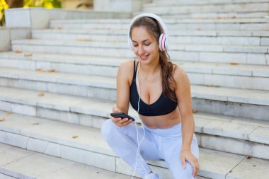 Eğitimden sonra dinlenme kavramı. Spor giyim sektöründe genç ve mutlu bir kadın akıllı telefon kullanıyor ve güneşli bir günde merdivenlerde otururken kulaklıkla müzik dinliyor.