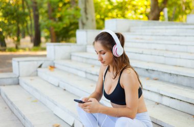 Eğitimden sonra dinlenme kavramı. Spor giyim sektöründe genç bir bayan güneşli bir günde merdivenlerde otururken akıllı telefon kullanır ve kulaklıkla müzik dinler.