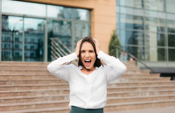 Young hysterical business woman screaming and holding her head her arms against the background of a business center