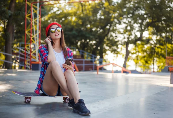 Young stylish woman dressed in youth clothes sits on a skateboard and listens to music on headphones at a skatepark. Summertime