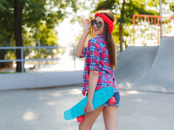 Parlak güneşli bir günde kaykay parkında elinde kaykay tutarken şık giyinmiş neşeli, duygusal genç bir kadın.