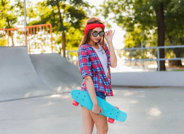 Parlak güneşli bir günde kaykay parkında elinde kaykay tutarken şık giyinmiş neşeli, duygusal genç bir kadın.