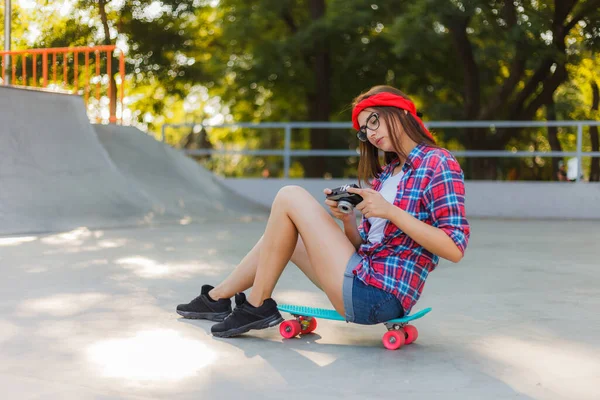 スタイリッシュな服の若い女性はスケートボードに座って 明るい晴れた日にスケートパークでレトロなカメラを使用しています 若者の概念 — ストック写真