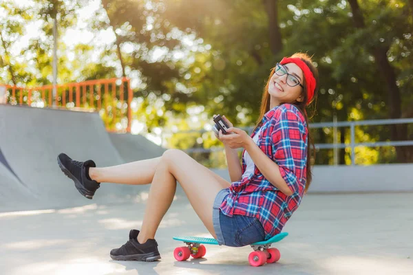 スタイリッシュな服の面白い若い女性はスケートボードに座って 明るい晴れた日にスケートパークでレトロなカメラを使用しています 若者の概念 — ストック写真