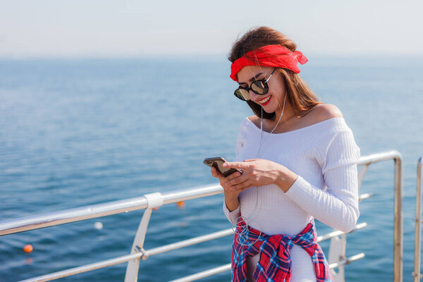 Stylishly dressed young woman listens to music in earphones and uses smartphone on beach against sea background. Street Fashion