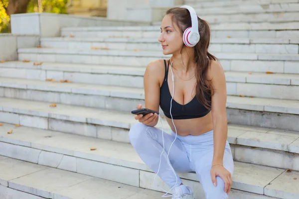 Eğitimden sonra dinlenme kavramı. Spor giyim sektöründe genç ve mutlu bir kadın akıllı telefon kullanıyor ve güneşli bir günde merdivenlerde otururken kulaklıkla müzik dinliyor.