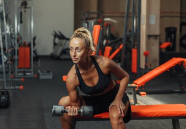 Spor salonundaki bir bankta otururken, genç bir fitness kadını biseps için halter kaldırmaya konsantre oluyor. Serbest ağırlık ile eğitim kavramı.