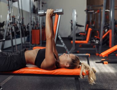 Spor kıyafetli zayıf bir kadın spor salonunda bir bankta uzanırken halterlerle ağırlık kaldırma egzersizi yapıyor. Serbest ağırlık çalıştırma konsepti. Fonksiyonel eğitim