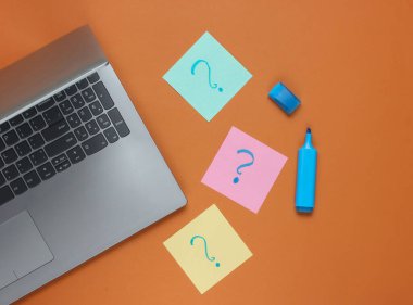 Laptop, üzerinde soru işaretleri ve kahverengi arka planda keçeli kalem olan not kağıtları. Üst görünüm