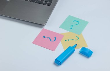 Laptop, beyaz arka planda soru işaretleri ve keçeli kalemle çizilmiş not kağıtları. Üst görünüm