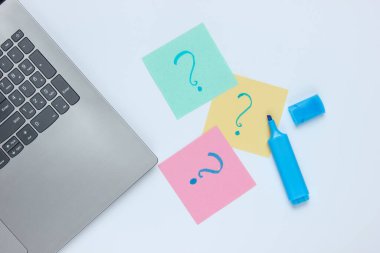 Laptop, beyaz arka planda soru işaretleri ve keçeli kalemle çizilmiş not kağıtları. Üst görünüm