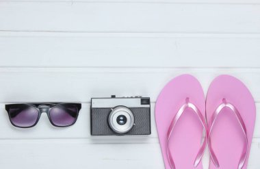 Plajdaki turizm aksesuarları. Plaj tatili Parmak arası terlikler, retro kamera, beyaz ahşap zeminde güneş gözlüğü. Üst görünüm
