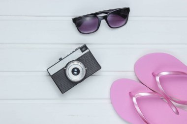 Plajdaki turizm aksesuarları. Plaj tatili Parmak arası terlikler, retro kamera, beyaz ahşap zeminde güneş gözlüğü. Üst görünüm
