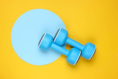Sarı arka planda, mavi pastel halkalı plastik halterler. Üst Manzara. Minimalist spor konsepti. Üst görünüm