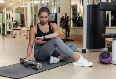 Sağlıklı yaşam tarzı kavramı. Genç sıska kadın antrenmandan sonra mindere oturmuş dinleniyor ve spor salonunda saat izliyor.