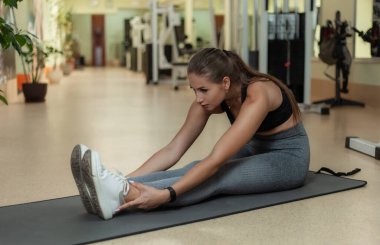 Genç ve çekici bir kadın spor salonunda paspasın üzerinde otururken bacak esnetme egzersizi yapıyor.