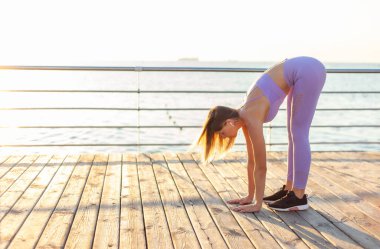 Sabah sporu. Eğitimden önce ısınıyorum. Spor kıyafetli genç bir kadın güneş doğarken sahilde bacak esnetiyor.